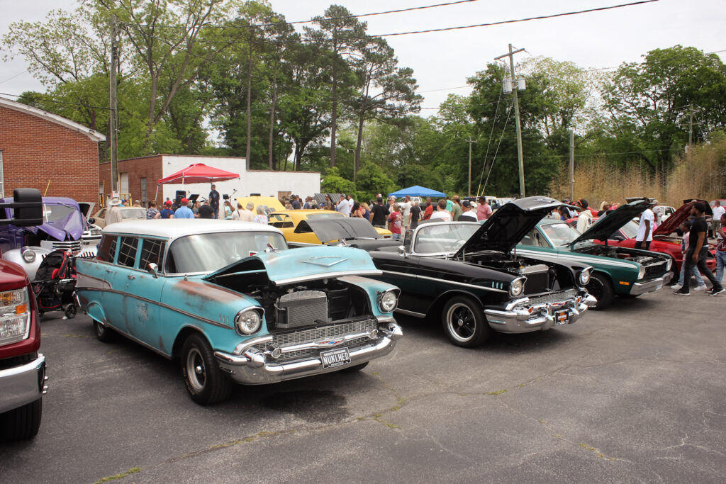 A lively outdoor car show featuring multiple vintage vehicles, including a weathered turquoise Chevrolet and a sleek black Chevrolet with hoods open for display. A crowd of people is gathered around, admiring the cars, with tents and booths set up in the background. The event takes place in a parking lot surrounded by trees and brick buildings.