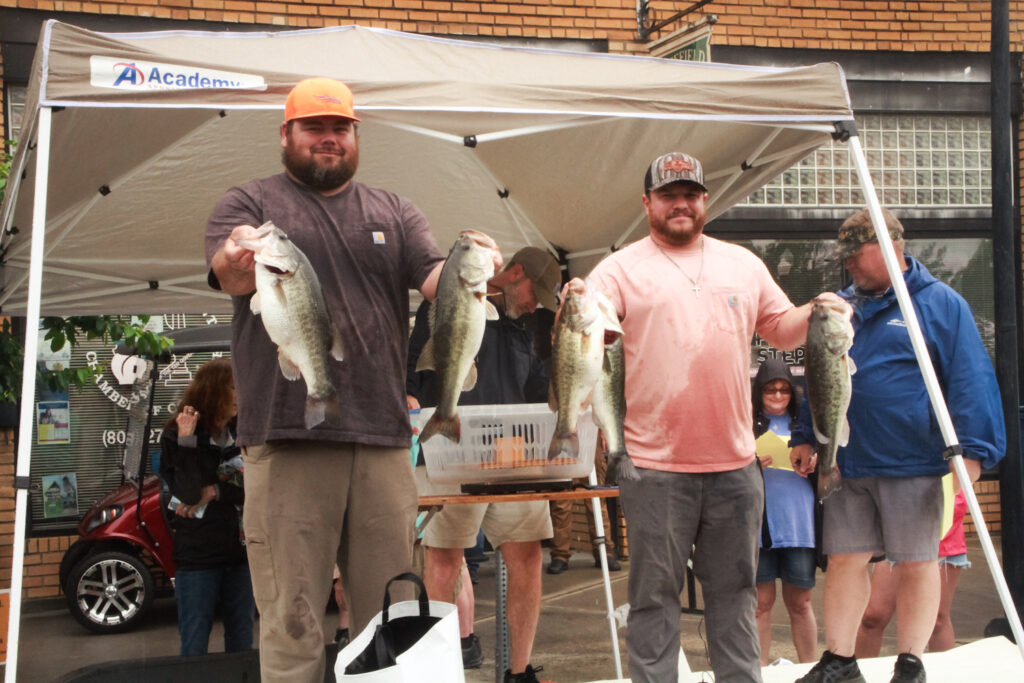 Two men stand under a canopy, each holding large bass fish they caught. They are smiling and appear to be participating in a fishing competition. A small crowd is visible in the background, with a table and weighing station set up behind them. One man wears a dark brown shirt and an orange cap, while the other wears a salmon-colored shirt and a camo cap.